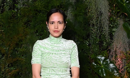 Anna Jewsbury at the British Vogue x Netflix Bafta party in London, May 2024: she has dark brown hair tied back to show the multiple pairs of ornate silver, gold and pearl earrings she is wearing; she wears a pale silvery-green high-necked and short-sleeved velvet top with a leaf-like pattern and is looking straight at the camera. She has dark brown eyes and is wearing pink lip gloss; she stands against a dark backdrop of trailing plants and leaves
