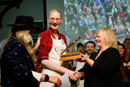 Gina Saarberg receives her golden spurtle prize. She is in her late 20s with fair hair tied back, and wears a red shirt and cream apron plus large spectacles.