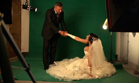 A woman in a white dress, glove and a veil sits on the floor of a photographic studio holding a hand out to a man in a suit who bends towards her