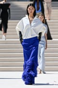 A fashion model walks down an outdoor runway wearing a structured white blouse with an oversized, sculptural silhouette and a deep V-neckline with long black gloves