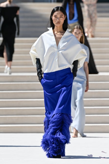 A fashion model walks down an outdoor runway wearing a structured white blouse with an oversized, sculptural silhouette and a deep V-neckline with long black gloves