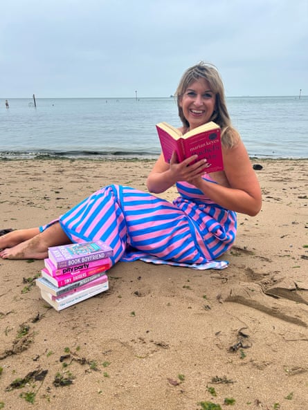 Daisy Buchanan on the beach in bright stripey dress reading a big pink book with a pile of brightly coloured books alongside - with sea in background