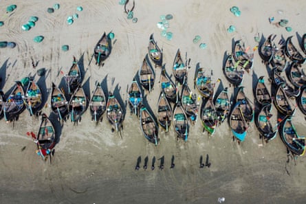 Aerial shot of shadows of seven people in a line passing 20 or so traditional wide-hulled fishing boats pulled up on a beach