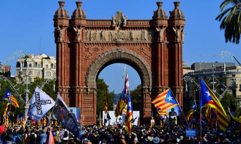 Carles Puigdemont delivers a speech on a stage in front of the Arc de Triomf, a tall and ornate brick monument, to a crowd of people waving flags qhiddrituiqhqinv