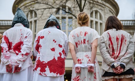 Four people with red paint splattered on their white clothes stand with their arms tied behind their backs during a Berlin protest against war crimes in Ukraine. One has 'stop war' written on their back.