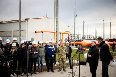 Viktor Orbán talks into a microphone while journalists wearing hardhats stand in front of him, with pipeline infrastructure in the background