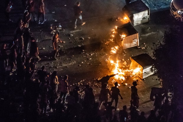 A crowd stand in a road behind a line of burning dumpsters