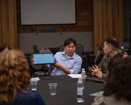 A man sitting at a table holds a small screen for others to see. Several people are also sitting at the table