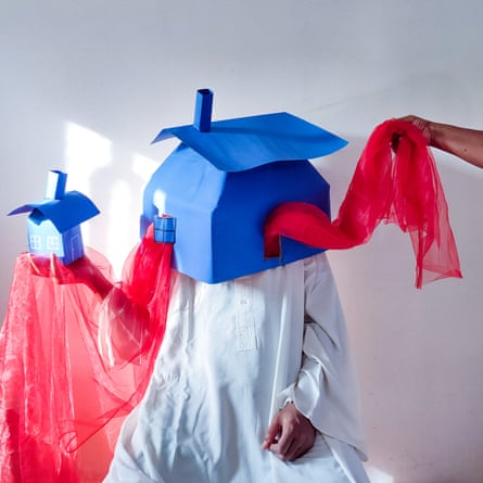 A person wearing a white robe has house made of blue cardboard on their head, and holds a smaller one in their right hand. Red fabric flows between them.