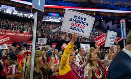 An Asian woman in a long-sleeved shirt, grinning, holds up a sign reading “Make America GREAT again!” in a crowd indoors at the Republic national convention