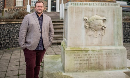 Terry Jermy stands beside a statue (plinth only seen) in Thetford town centre. He is in his late 30s and wears a light brown jacket over a dark patterned shirt and dark red jeans.