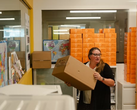 a woman holds a box with stacks of orange boxes behind her