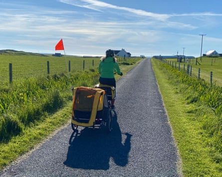 woman cycles away down a road pulling a child trailer amid a view of fields