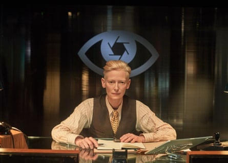 A woman with short blonde hair looks serious while sitting at a desk with a large logo of an eye behind her.