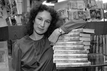 A black and white photograph of Margaret Atwood in a bookshop with her left arm leaning on a tall pile of her one of her books