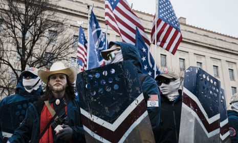 Thomas Ryan Rousseau, founder of white supremacist group Patriot Front, wears a white cowboy hat as he stands with other members of the group, who are wearing dark sunglasses and white masks that cover their faces below the nose. The group wears jackets with the Patriot Front insignia and carry flags bearing the Patriot Front logo. One member carries an upside-down US flag.