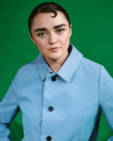 Maisie Williams pictured from the waist up with her hands on her hips, looking at the camera, wearing a pale-blue mac with black buttons, against a green background