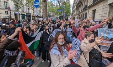 Young people standing on a street, many wearing face masks and keffiyeh scarves, some holding a Palestinian flag