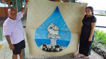A man and a woman hold up a woven grass mat with an image of a fist emerging from a nuclear mushroom cloud