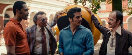 Bunch of men stood around a guy in a blue shirt, all wearing 70s clothing, trying to intimidate him