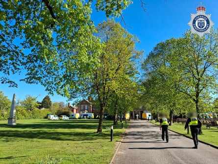Police officers walk along a tree-lined road in parkland towards a large building.