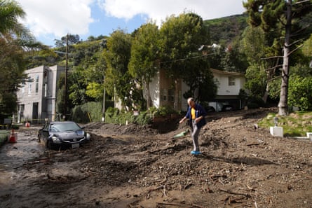 A pretty, green, urban street completely covered in mud, with a man walking on top of it and a half-buried car at the edge.