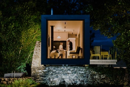 A man and woman inside a room at Barneys Ruins, seen from outside at night through a window taking up the whole of one end of a shipping container.