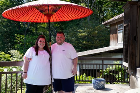 Marisa and Russell Crossan, both wearing white T-shirts and black shorts, stand under a red pleated umbrella on a visit to Japan in Around the World in 80 Weighs