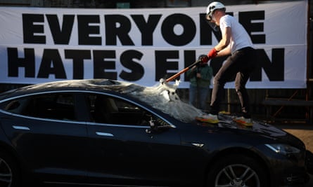 A man wearing a protective helmet stands on the bonnet of the car and smashes the windscreen with a sledgehammer. A banner behind the car says ‘everyone hates Elon’
