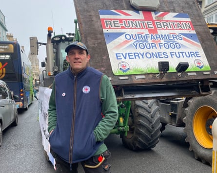 Derek Pearce stands with his hands in his jacket pockets. Behind him, a sign on a tractor reads 're-united Brain. Our farmers, your food, everyone's future'