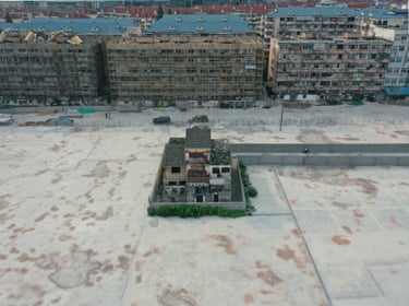 A solitary house surrounded by fencing with a walkway to it, housing estates in the background