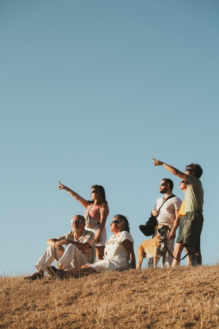A group of people wearing eclipse glasses sit and stand on a hill, pointing at the sky