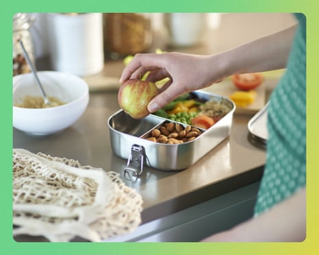 A hand places an apple into a plastic-free lunchbox filled with almonds, peas, and vegetables