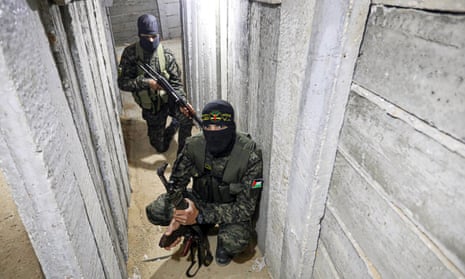 Two militants wearing masks and holding guns crouch on a sandy floor between concrete walls