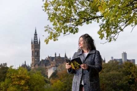 Prof Hester Parr stands in Kelvingrove Park, Glasgow, with a tall spired building in the background; she has curly shoulder-length dark grey hair and wears a grey tweed coat and red lipstick. She is holding a booklet and smiling.