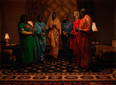 Six women in colourful embroidered robes stand talking together in a dimly lit room