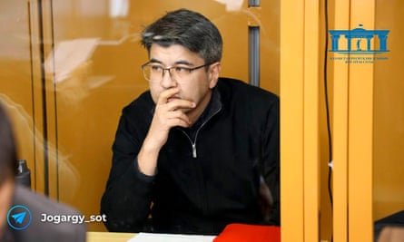 A man in glasses sits in a defendant’s cage in a court room