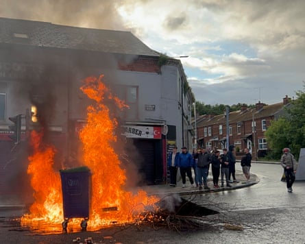 Wheelie bins ablaze on the street, a small group stands nearby.