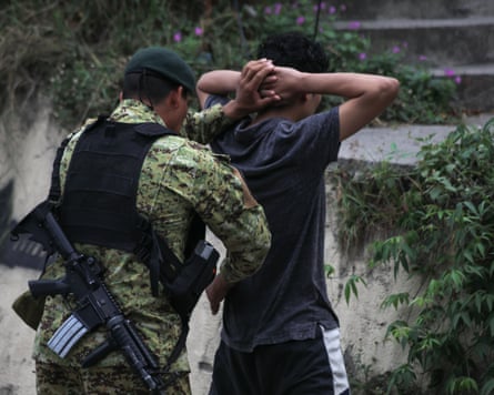 A young man stands with his hands on his head. Another man wearing fatigues and with a gun slung over his shoulder stands behind him holding his hands with one hand and appearing to search him with the other