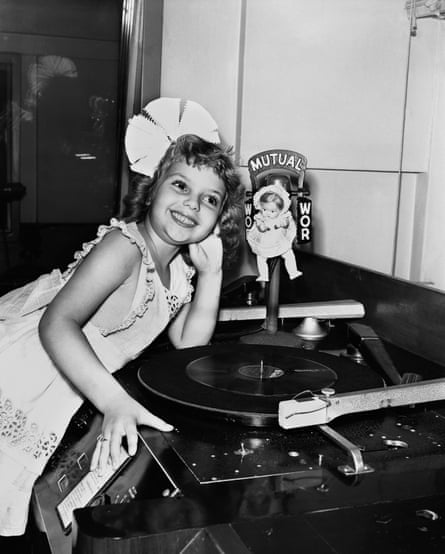 A black-and-white photo of a beaming little girl with a white hairbow as big as her head, next to a record player.