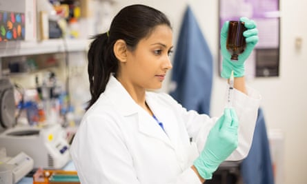 Stock image of young south Asian woman with long black hair pulled back wearing white lab coat and green gloves, holding lab equipment.
