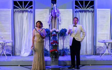 A man in a white evening jacket, bow tie and black trousers and a woman in a vintage ballgown stand in front of a set that looks like the exterior of a house with long french windows and a verandah.