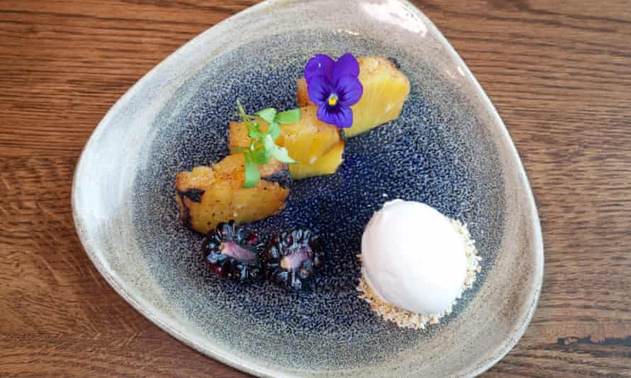 A blue/grey triangular plate with three chunks of pineapple with edible flowers on top, and a scoop of white sorbet