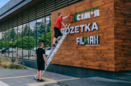 A man stands on a ladder removing a sign from exterior of a supermarket, as another man looks on