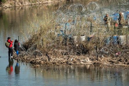 A couple wades in a river to a bank covered in barbed wire while military men look on.