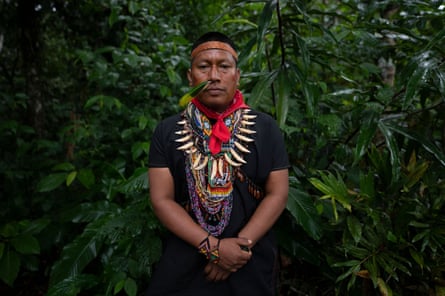 An Indigenous man with his face lightly painted and in traditional dress adorned with necklaces of beads and large animal teeth and a green feather coming out of his nose