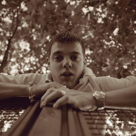A young Mike Skinner leaning over a fence, looking down at a camera on the floor, with the leaves of trees above him