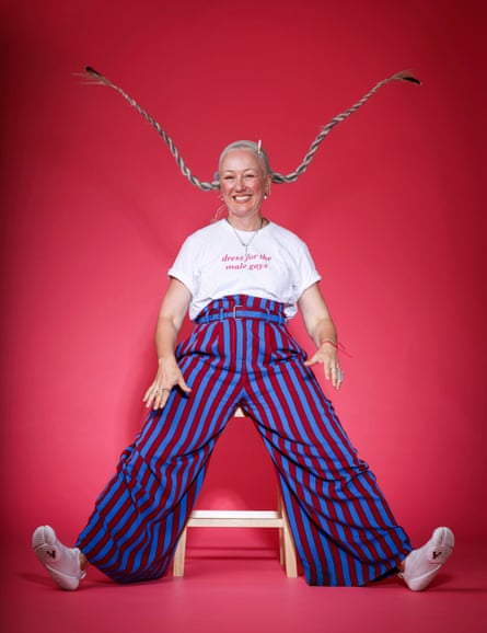 Anouchka Grose sitting on a tall white stool against a red background with her legs stretch out to the side, wearing a white T-shirt with “dress for the male gays” on it and high-waisted’ belted baggy trousers with blue and red vertical stripes on, white trainers and a silver necklace, with her silver hair in very long plaits that are sticking up in the air and a cigarette on her head