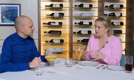 A bald man wearing a bright blue patterned jumper, and a blond woman wearing a pink cardigan, sitting at a restaurant table talking