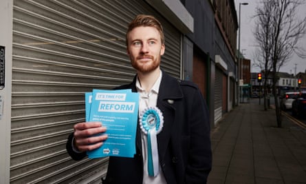 Jamie Gregory holds up blue Reform leaflets whilst wearing a blue Reform rosette